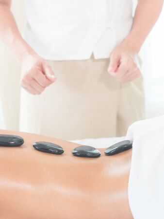 An expert masseuse at a spa salon puts hot stone on the back of an Asian woman to relieve tension...