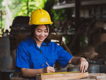 Woman working in mechanic uniform using measuring tools to cut wood sheets in factoryの写真素材