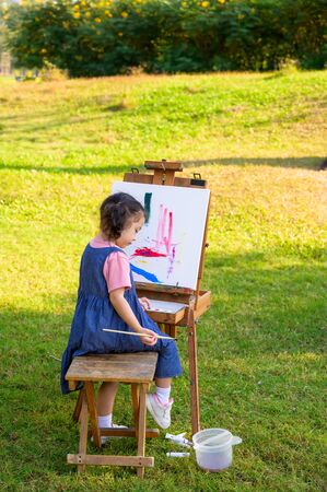A little girl is sitting on the wooden bench and painted on the canvas placed on a drawing stand, Be a part of learning outside of the school in the nature parkの写真素材