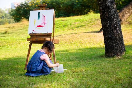 A little girl is sitting on the grass and painted on the canvas placed on a drawing stand, Be a part of learning outside of the school in the nature parkの写真素材