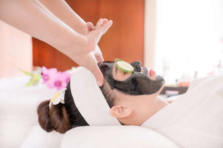 A beautiful Asian woman uses spa mud and cucumber for facial treatment, along with a massage by a professional masseuse in the spa salonの写真素材