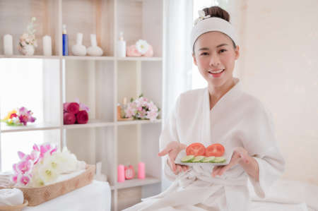A beautiful Asian woman prepares to use tomatoes and cucumbers for facial care, along with a massage by a professional masseuse in a spa salonの写真素材