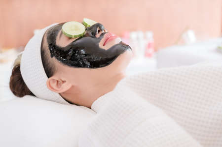 A beautiful Asian woman uses spa mud and cucumber for facial treatment, along with a massage by a professional masseuse in the spa salonの写真素材