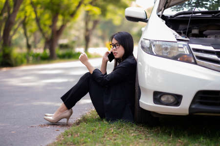 A young Asian woman is calling her service technician to fix a broken car on the side of the road, Due to an accident The engine makes it impossible to continue drivingの写真素材
