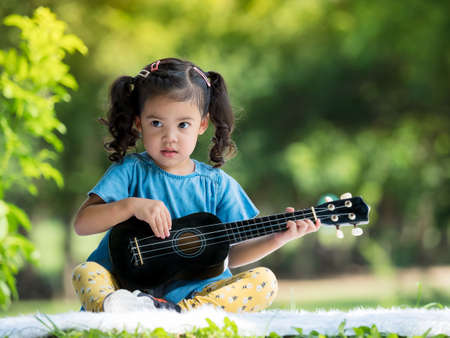 Asian little girl sitting  on the carpet, Relax and playing ukulele outside of school to enjoy in the nature parkの写真素材