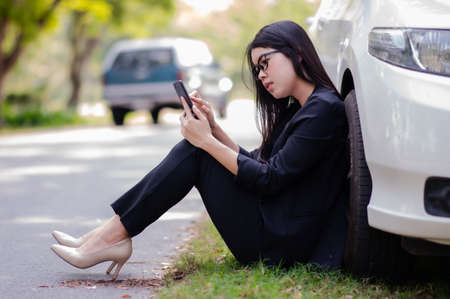 A young Asian woman is calling her service technician to fix a broken car on the side of the road, Due to an accident The engine makes it impossible to continue drivingの写真素材