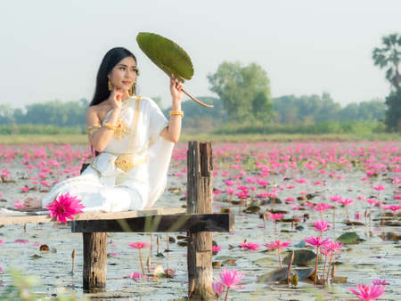 An elegant Thai woman wearing traditional Thai clothes adorned with gold ornaments carrying lotus flowers leaf collected from a lotus fieldの写真素材