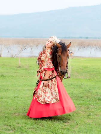 A beautiful western woman in a beautiful dress stands with her horse in the lake meadowの写真素材