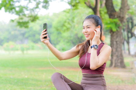Pretty Asian woman takes a break to listen to music on her mobile phone after exercising in the park, It is a way of life for relaxation and good healthの写真素材