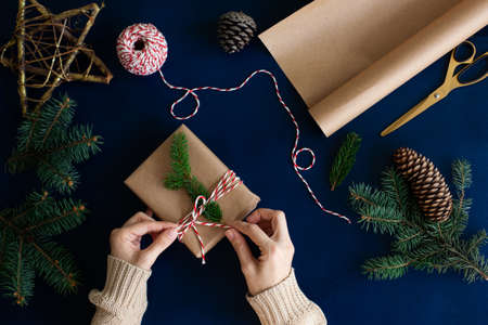 woman wraps a handmade eco style christmas present in kraft paper and natural spruce branches on a...