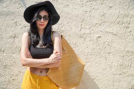 Stylish young woman in hat and sunglasses with straw bag, rough cement wall background.handbag on...