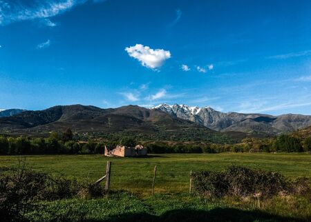 Ruined house in the middle of a green meadow at the foot of a snowy mountain and a blue sky with white clouds in Candeleda, Avilaの写真素材