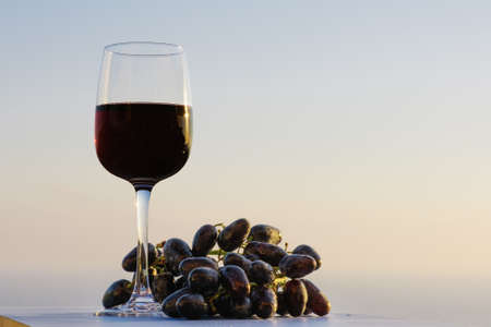 Wineglass and branch of black grapes on the sunset sky background. Balcony view. Red wine on the sea ocean background.の写真素材