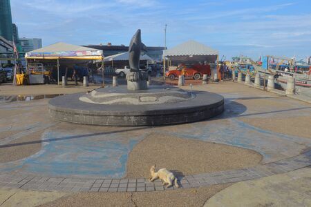 Kota Kinabalu, Sabah/Malaysia - December 21 2019 : A cat laying down on the pavement near a dolphin sculpture at the square of Kota Kinabalu Waterfront in the city of Kota Kinabalu, Malaysia.のeditorial素材