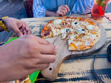 Chicken pizza with sausage and mushroom topping served on a wooden cutting board, placed on a wooden table at a beachside restaurant.の写真素材