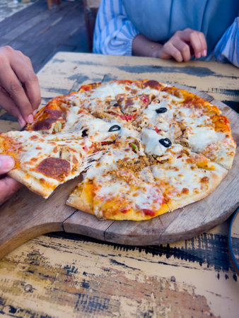 Chicken pizza with sausage and mushroom topping served on a wooden cutting board, placed on a wooden table at a beachside restaurant.の写真素材