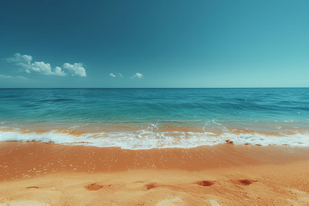A tranquil tropical beach scene featuring calm turquoise ocean waves lapping onto golden sand under a vast, clear blue sky with scattered white clouds.の素材