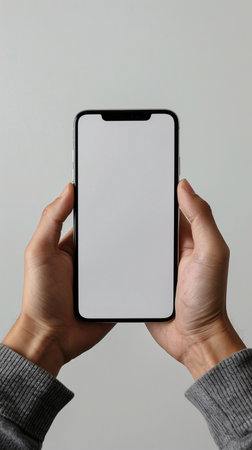 A close-up shot of two hands holding a contemporary smartphone with a white screen against a neutral background.の素材