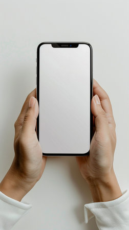 Top down shot of two hands holding a modern smartphone displaying a blank white screen, isolated on a light background.の素材