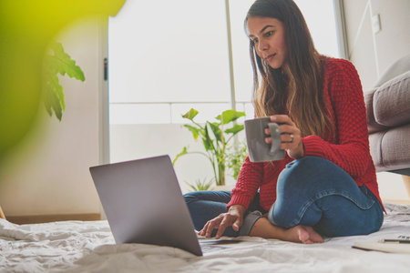 Portrait of a Latin woman drinking hot coffee and using laptop, watching video, free space. Sitting on the couch and relaxing in your cozy home. Concept creative work at home. Selective focus.の写真素材
