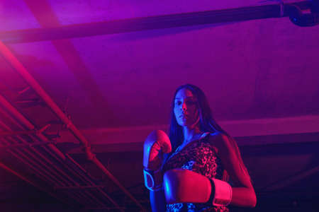 Beautiful female boxer training, red boxing gloves, ring with red and blue lights, industrial gym....