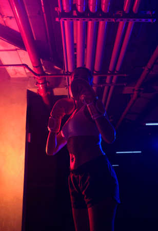 Beautiful female boxer training, red boxing gloves, ring with red and blue lights, industrial gym, neon lights. selective focusの写真素材