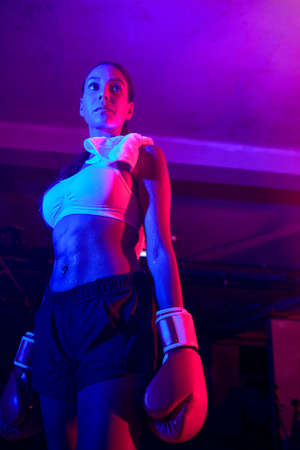 Beautiful female boxer training, red boxing gloves, ring with red and blue lights, industrial gym,...