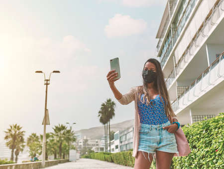 Young blond woman in red suit and with protective face mask taking a selfie photo near luxury...