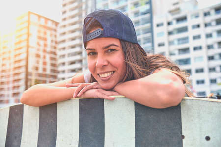 Beautiful young woman in cap is sitting at skatepark on the ramp with her longboard.の写真素材