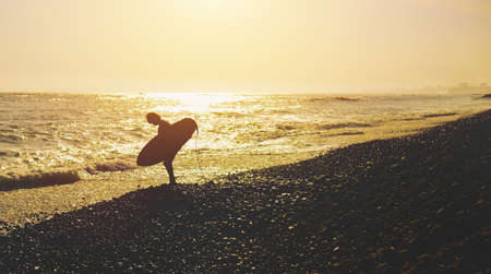 Unrecognizable silhouette of male surfer with his surfboard walking away. Backlight during sunset with dramatic sky and reflection.の写真素材