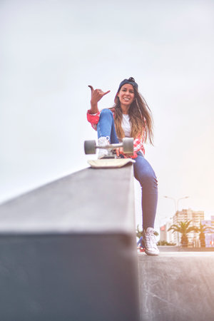 Beautiful young woman in cap is sitting at skatepark on the ramp with her longboard.の写真素材