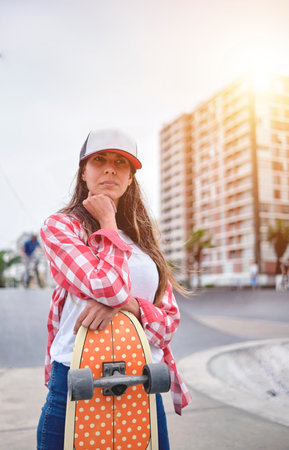 Beautiful young woman in cap is sitting at skatepark on the ramp with her longboard.の写真素材