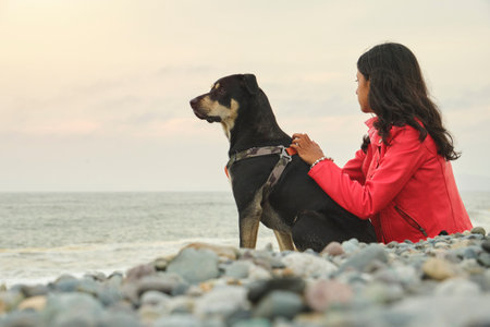 A woman sits on the beach with her dog. The dog is wearing a harness and he is enjoying the beach....