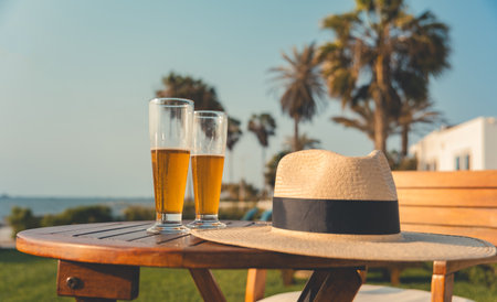 A table with two glasses of beer and a straw hat on itの写真素材