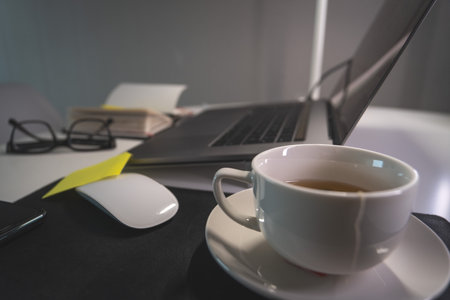 A white coffee cup sits on a table next to a laptopの写真素材