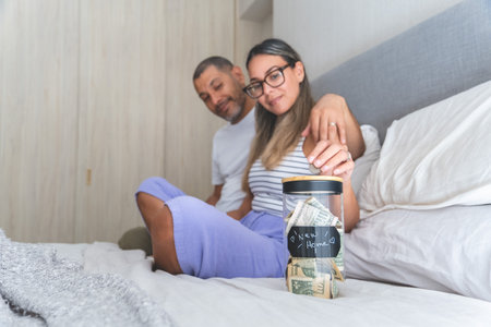 A man and woman are sitting on a bed and the woman is putting money into a jar. New home concept.の写真素材