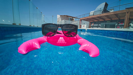 Pink inflatable toy crab and sunglass floating in blue swimming pool during summer vacation in...