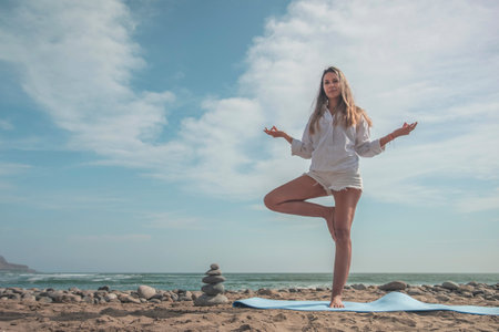 Yoga instructor practicing balancing tree pose on beach yoga mat during summer meditation timeの写真素材