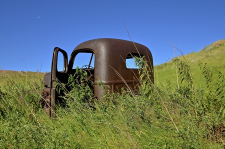 The rusty deserted cab of an old pickup is surrounded by long weeds in a hilly environmentの写真素材