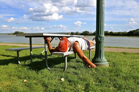 An identified man who is sleeping on a picnic table bench by a large river indicates patriotism as defined by head gear.の写真素材