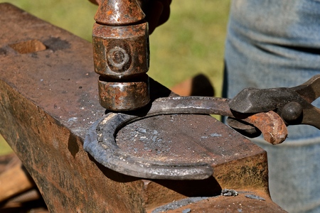 A heavy metal hammer strikes a horseshoe held by a tongs on top of an anvilの写真素材