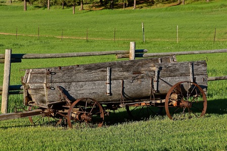 An old weathered wood wagon with steel wheel serves as a yard decoration against a wood railing fence.の写真素材
