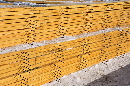 A stacked pile of pre-made rafters with protruding screws lie in the cold snow.