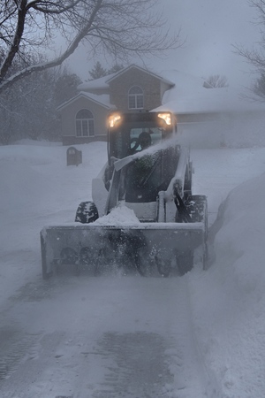 A skid steer with a an attached snow blower clears a path through the deep snow  during a blizzard.