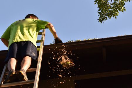An unidentified man stands on an extension ladder cleaning the eaves and gutters of a two story houseの写真素材