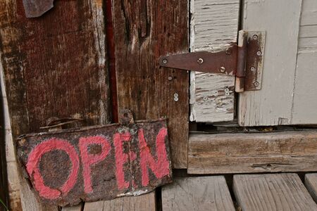 An old wooden and rustic door with a rusty hinge has a sign painted in bright lettering stating they are open.の写真素材