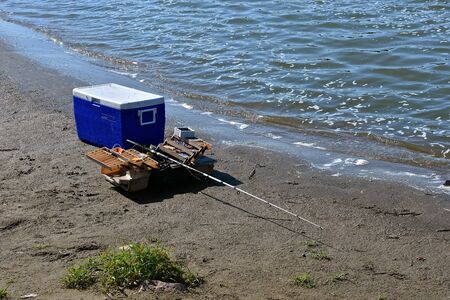 A fisherman leaves a cooler, rod and reel, and tackle box along the shore of a lake or river.の写真素材
