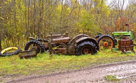 Old discarded tractors are lined up between a woods and a very muddy roadの写真素材
