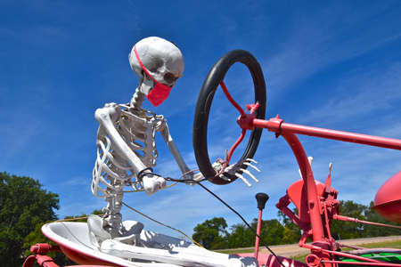 A skeleton with a Covid-19 mask is steering an old red tractor.のeditorial素材