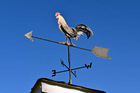 A wind vane on top of a rooftop displays a rooster outlined against a clear blue skyの写真素材
